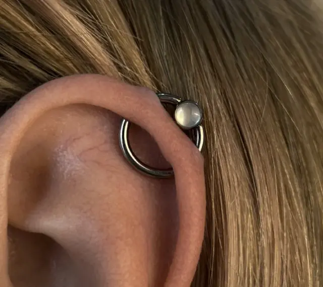 Oryginalny kolczyk orbital piercing z białym kamieniem na uchu, otoczony blond włosami.
