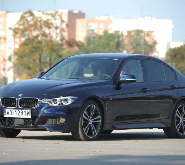 Ciemnoniebieskie BMW 318d z polskimi tablicami rejestracyjnymi stoi na tle budynków i drzew.