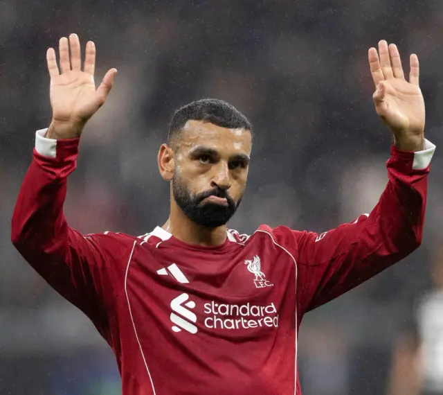Mohamed Salah w czerwonej koszulce Liverpoolu, z uniesionymi dłońmi, pozdrawia kibiców. Widać logo klubu i sponsora.