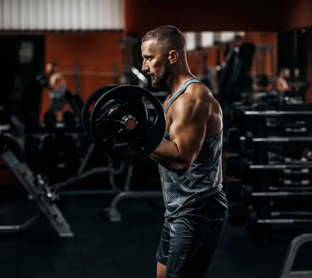 Mężczyzna ćwiczy biceps z ciężarem. W tle siłownia, sprzęt i inny ćwiczący.