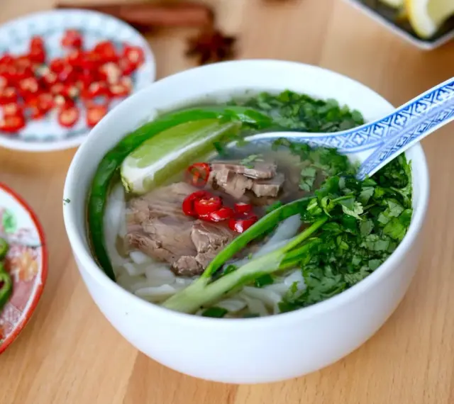 Aromatyczna zupa pho z wołowiną, makaronem ryżowym, świeżą kolendrą, chili i limonką. Obok talerzyki z dodatkami.