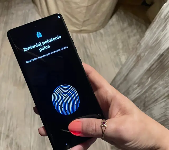 Dłoń z telefonem, na ekranie skaner linii papilarnych i komunikat "Zmieniaj położenie palca". To jak próba ucieczki z cyfrowego więzienia, niczym **jailbreak**.