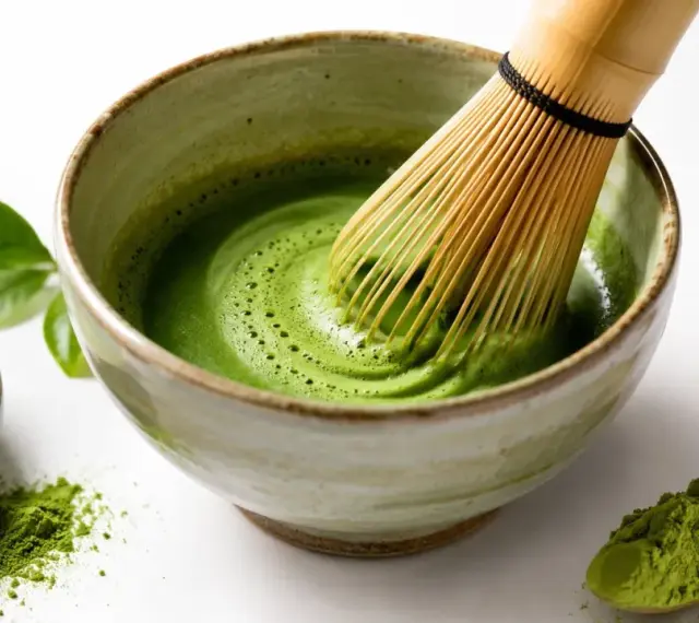 Tradycyjne matcha przygotowanie: bambusowa miotełka miesza zielony proszek z wodą w miseczce, tworząc piankę. Obok mała miseczka z proszkiem i drewniana łyżeczka.