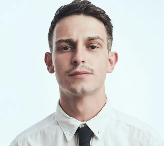 Młody mężczyzna, Vito Bambino, w białej koszuli i czarnym krawacie. Jego wzrost jest imponujący, a spojrzenie intensywne.
