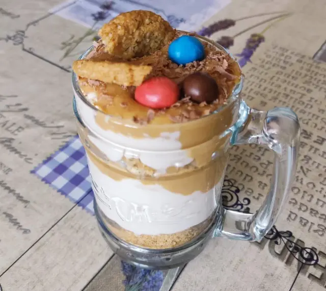 Warstwowy deser banoffee w szklance, ozdobiony ciasteczkiem, cukierkami i wiórkami czekolady.