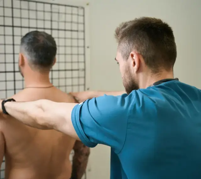 Therapeut führt krankengymnastik schulter Übungen durch. Ein Mann steht vor einem Gitter, während ein Therapeut seine Schultern berührt.