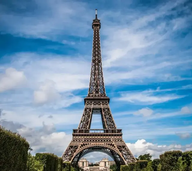 Majestatyczna wieża Eiffel w Paryżu, górująca nad zielonymi krzewami i błękitnym niebem z chmurami.