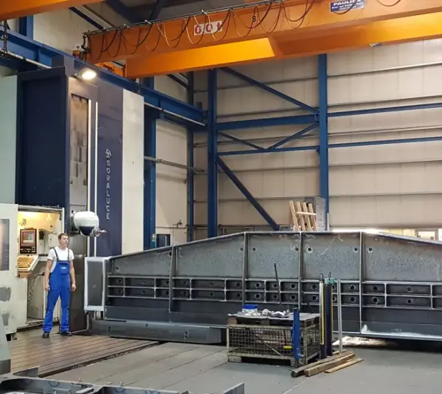 Ein junger Zerspanungsmechaniker in blauer Latzhose steht vor einer großen Soraluce-Maschine in einer Werkshalle. Daneben liegen massive Metallteile.