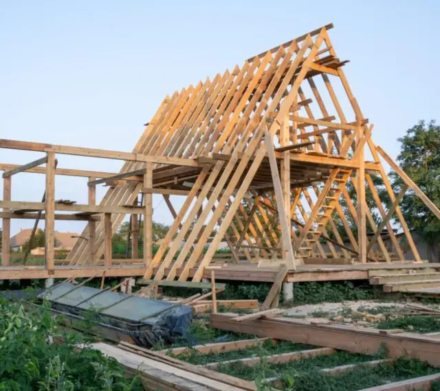 Drewniana konstrukcja domu w budowie. Widoczny szkielet dachu i ścian, schody. Fundament punktowy pod domek drewniany jest już gotowy.