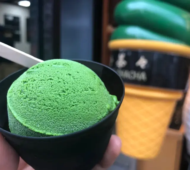 Zielone, japońskie lody matcha w czarnym kubeczku, z drewnianą łyżeczką. W tle wielki, żółty rożek z lodami.