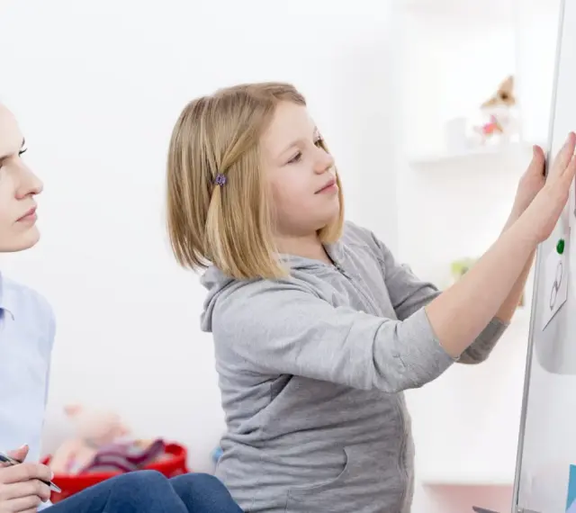 Terapeutaka z piórem w dłoni obserwuje dziewczynkę przyczepiającą rysunki do tablicy, analizując jej słabe strony.