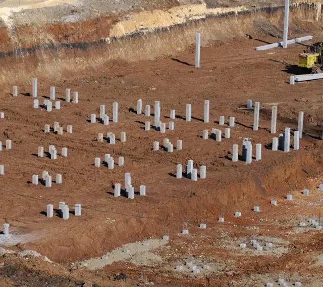 Plac budowy z licznymi betonowymi słupami, tworzącymi fundament pośredni. W tle maszyna budowlana.