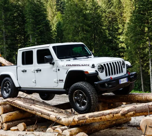 Biały jeep pickup Rubicon pokonuje przeszkodę z bali drewna w lesie.