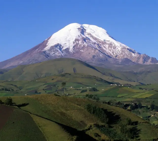 Pokryty śniegiem wulkan Chimborazo, jeden z najwyższych gór świata, góruje nad zielonymi, pofałdowanymi wzgórzami z polami uprawnymi.