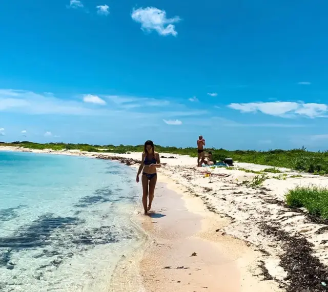 Kobieta spaceruje po piaszczystej plaży nad turkusową wodą. W tle widać innych ludzi i bujną zieleń. To idealne miejsce na wypoczynek nad Zatoką Meksykańską.
