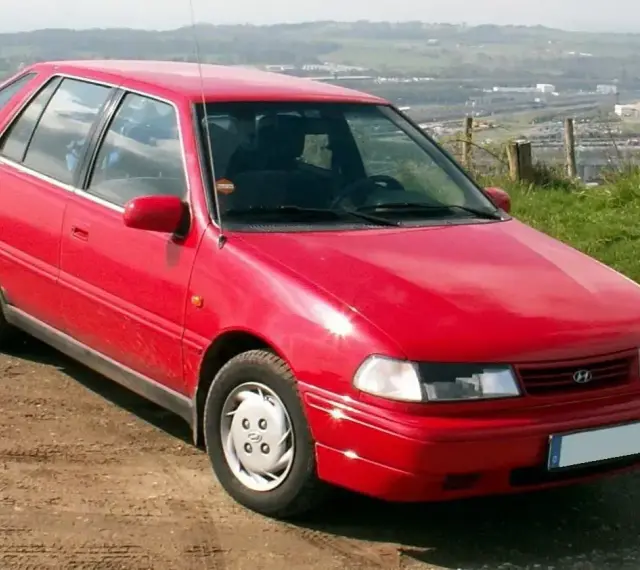 Czerwony Hyundai Pony z lat 90. stoi na polnej drodze, z widokiem na dolinę i miasto w tle.