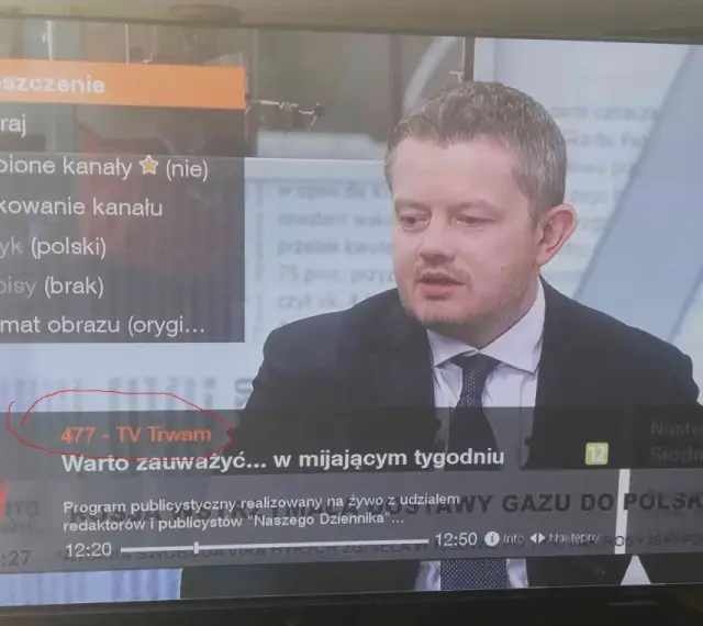 Mężczyzna w garniturze na ekranie telewizora, na którym widać menu z opcją "477 - TV Trwam".
