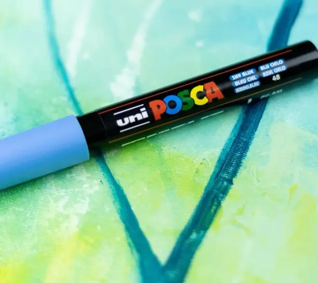 En ljusblå Posca-tuschpenna ligger på en abstrakt målning i gröna och blå nyanser. Perfekt för att måla med tuschpennor.