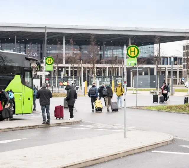 Ludzie z bagażami wsiadają do zielonego autokaru przed nowoczesnym budynkiem, przypominającym lotniska w Berlinie.
