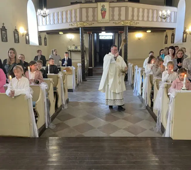 Uroczyste odnowienie przyrzeczeń chrzcielnych w kościele. Dzieci z zapalonymi świecami siedzą w ławkach, a ksiądz błogosławi zgromadzonych.