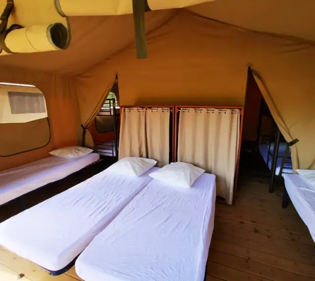 Intérieur d'une chambre de colonie de vacances avec plusieurs lits simples, draps blancs et oreillers. Des rideaux séparent les espaces.