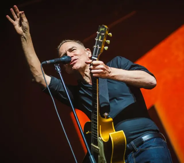 Bryan Adams na koncercie, w czarnej koszuli, z gitarą i mikrofonem, wznosi rękę w geście pozdrowienia.