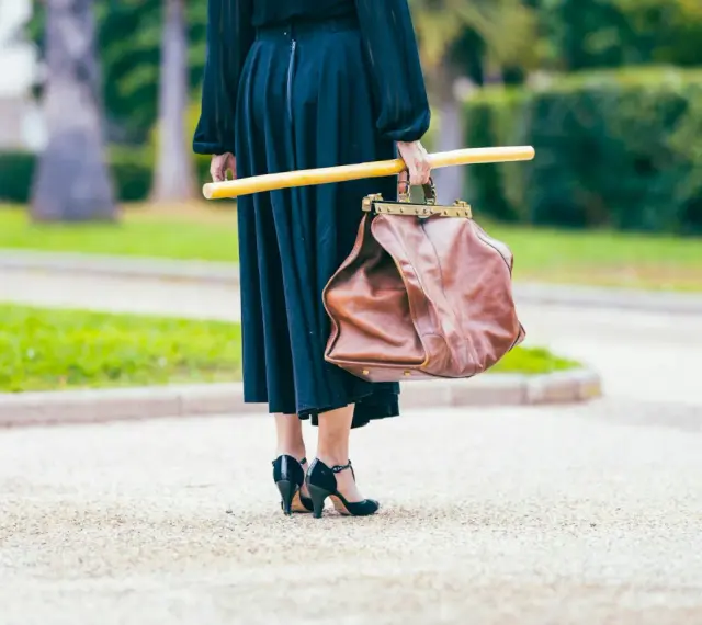 Elegancka kobieta w czarnej sukni i czarnych szpilkach, niosąca brązową torbę i żółty kij. Uosobienie francuskiej mody.