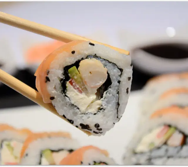 Chopsticks trzymają sushi z krewetkami, awokado i kremowym serkiem. W tle widać więcej roladek sushi.