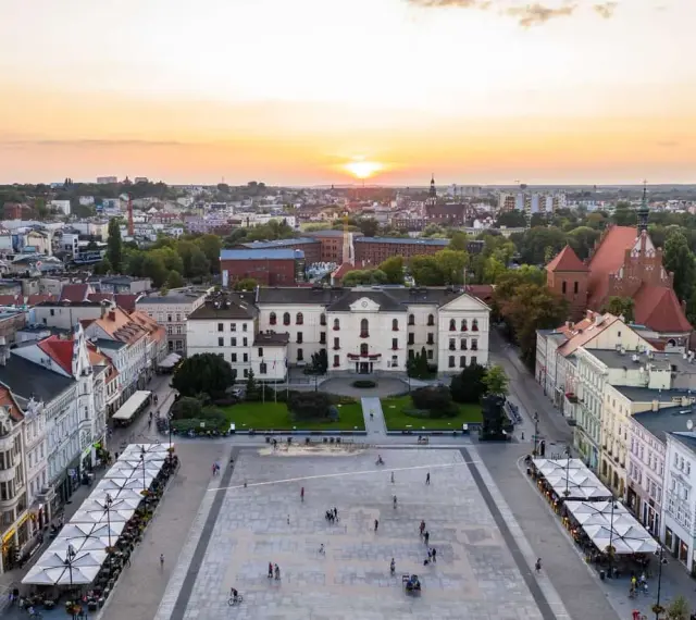 Zachód słońca nad rynkiem, gdzie widać piękną pierzeję kamienic i ratusz. Ludzie spacerują po placu, ciesząc się ciepłym wieczorem.