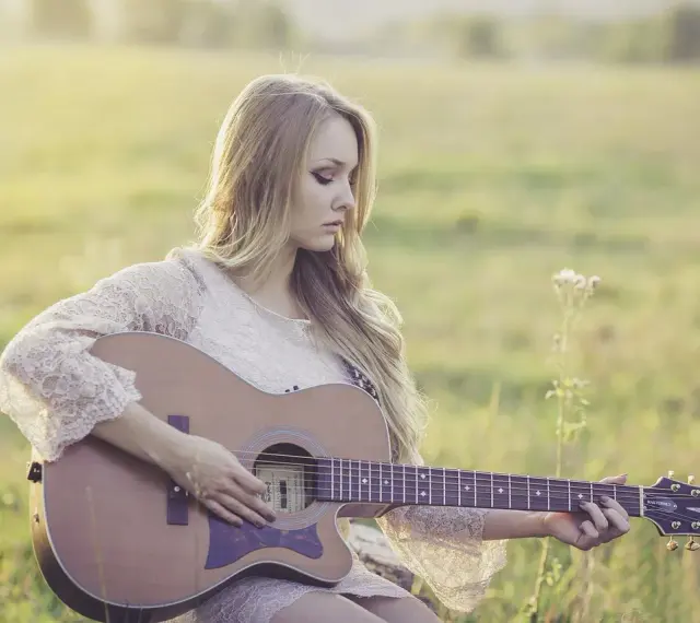 Blondynka w koronkowej sukience gra na gitarze w słonecznym polu. To jak remaster starych wspomnień, pełen ciepła i nostalgii.