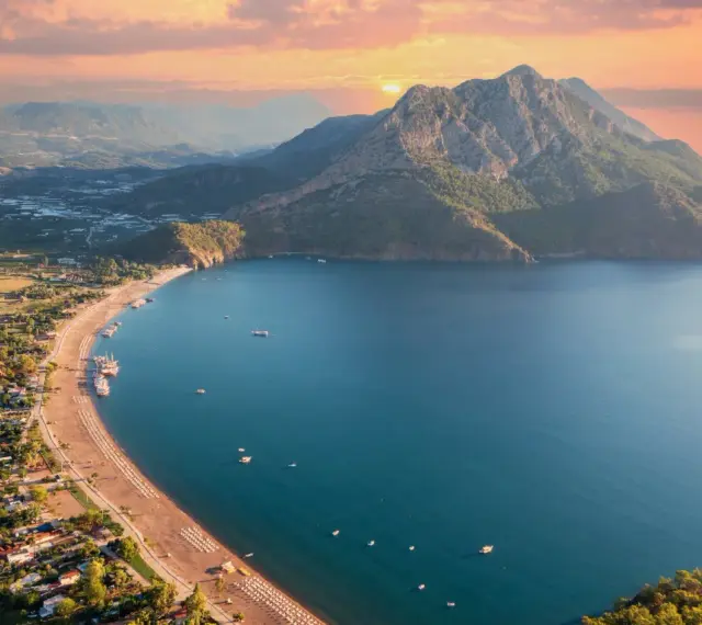 Malownicze wybrzeże Turcji o zachodzie słońca. Góry schodzą do turkusowego morza, a plaża z leżakami i łodziami zaprasza do odpoczynku.