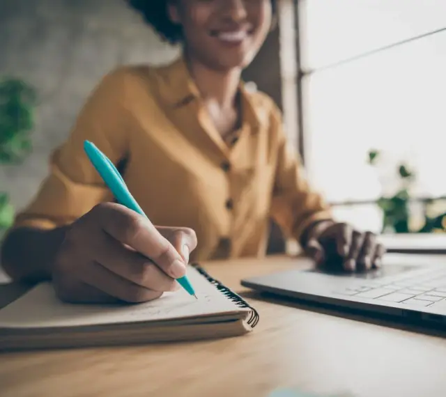 Frau schreibt mit einem blauen Stift Notizen in ein Notizbuch, während sie auf ihrem Laptop tippt. Sie erstellt ein Protokoll ihrer Gedanken.