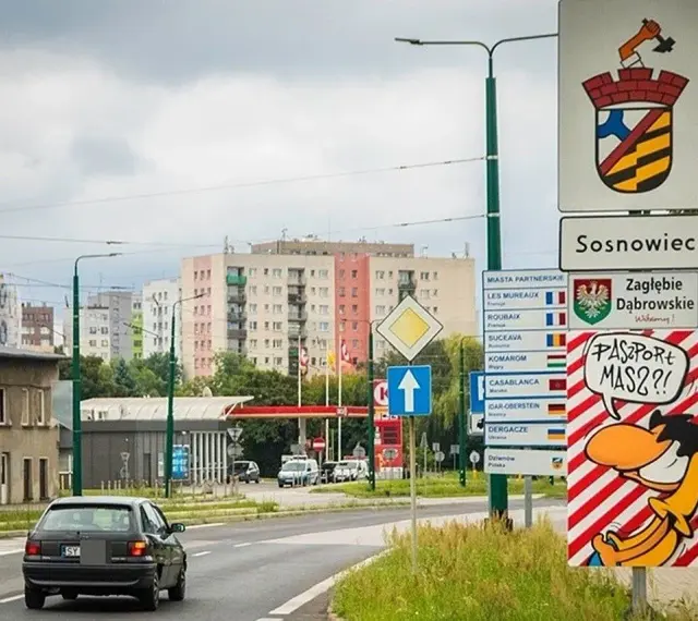 Znak drogowy "Sosnowiec" i humorystyczny napis "Paszport masz?!" przy wjeździe do miasta. W tle bloki mieszkalne.