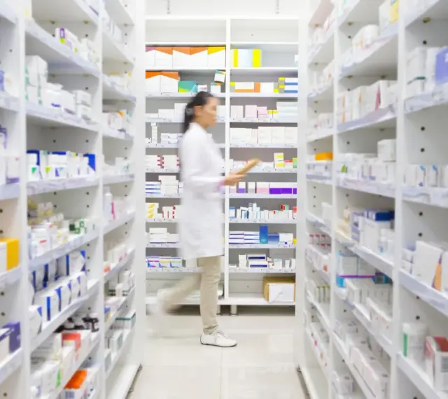Farmaceutka w białym fartuchu idzie między regałami pełnymi leków, szukając amoksycyliny.
