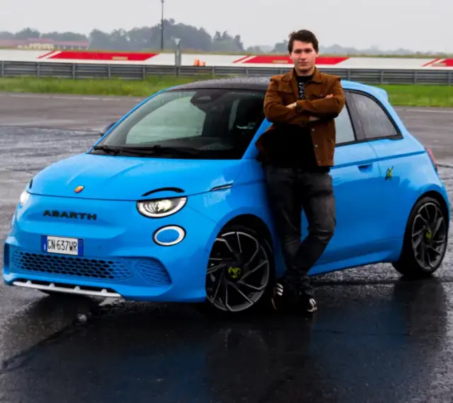 Mężczyzna opiera się o jaskrawo niebieski Abarth 500e na mokrym torze wyścigowym.