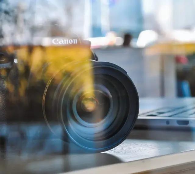 Winieta fotografia: obiektyw Canon na tle laptopa, odbicia tworzą artystyczny efekt.