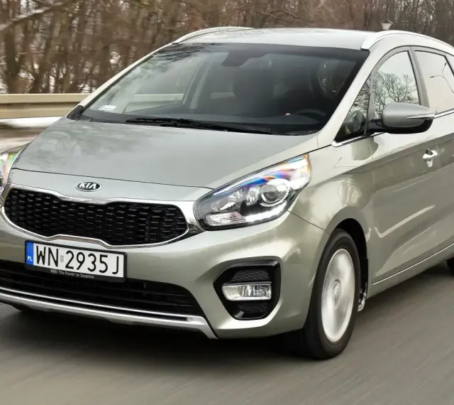 Srebrny Kia Carens, typowy MPV, jedzie po drodze. To auto rodzinne, idealne na wycieczki.