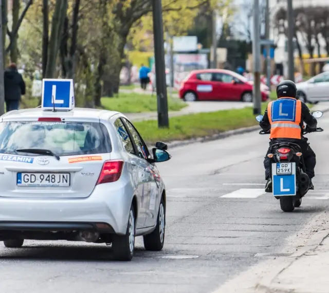 Srebrny samochód z literą "L" na dachu i motocyklista w kamizelce odblaskowej, obaj z literą "L" z tyłu, poruszają się po drodze. To kategoria a nauki jazdy.