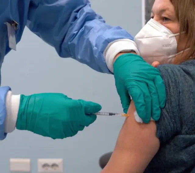 Osoba w niebieskim fartuchu i zielonych rękawiczkach podaje zastrzyk. Kobieta w masce KN95 otrzymuje trzecią dawkę szczepionki.