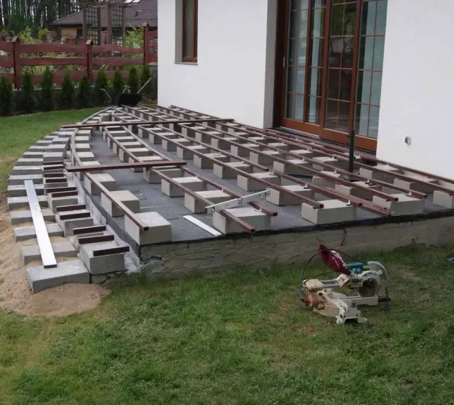 Budowa tarasu kompozytowego: betonowe bloczki podtrzymują legary, tworząc solidną konstrukcję. Widać narzędzia budowlane i materiały.