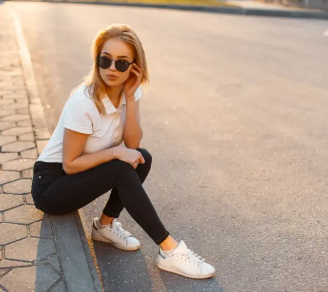Blondynka w czarnych jeansach i białej koszulce polo, siedząca na chodniku. Stylowa inspiracja, jak nosić koszulka polo damska na co dzień.