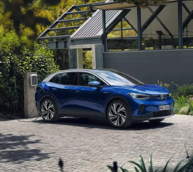 Niebieski Volkswagen ID.4 ładuje się przy domu, otoczony zielenią.
