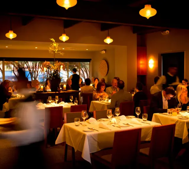 Ciemne, nastrojowe wnętrze restauracji. Goście jedzą przy stołach, kelnerzy krążą. Złe oświetlenie zdjęć jedzenia może zniechęcić gości do publikacji w social mediach.