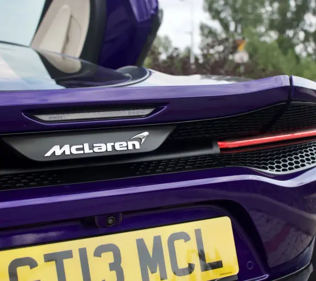 Fioletowy McLaren GT z otwartymi drzwiami, widok na tył z logo i tablicą rejestracyjną.