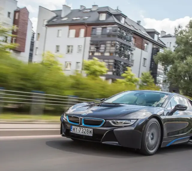 Czarny BMW i8 pędzi ulicą, mijając nowoczesne budynki i zielone drzewa.