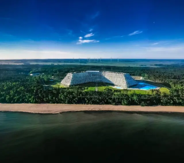 Hotel Gołębiewski w Pobierowie, otoczony lasem, z basenem i widokiem na morze. W oddali widać farmę wiatrową.