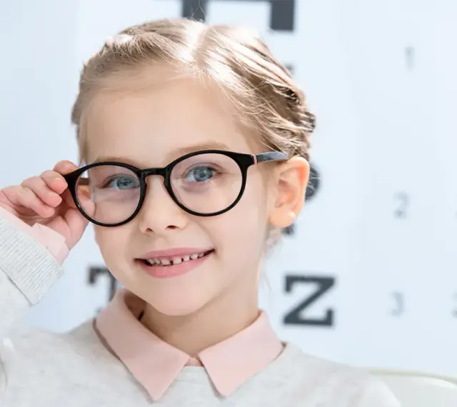 Uśmiechnięta dziewczynka w okularach, z tablicą optometryczną w tle. Okulary nosi się od jakiej wady, by widzieć wyraźnie.