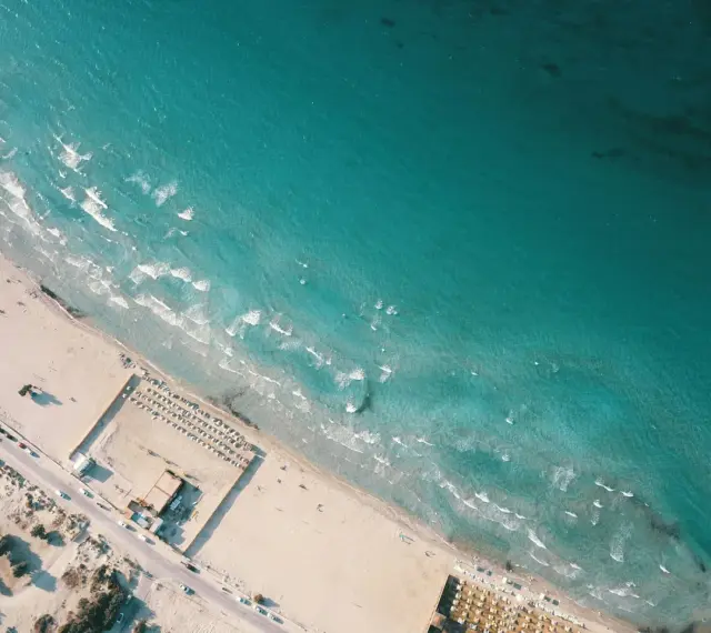 Izmir nad Morzem Egejskim. Piękna, piaszczysta plaża z turkusową wodą i falami. W oddali widać leżaki i parasole.