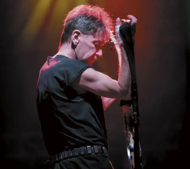 Muzyk z zespołu KSU z gitarą na scenie, w czarnej koszulce i z charakterystyczną fryzurą.