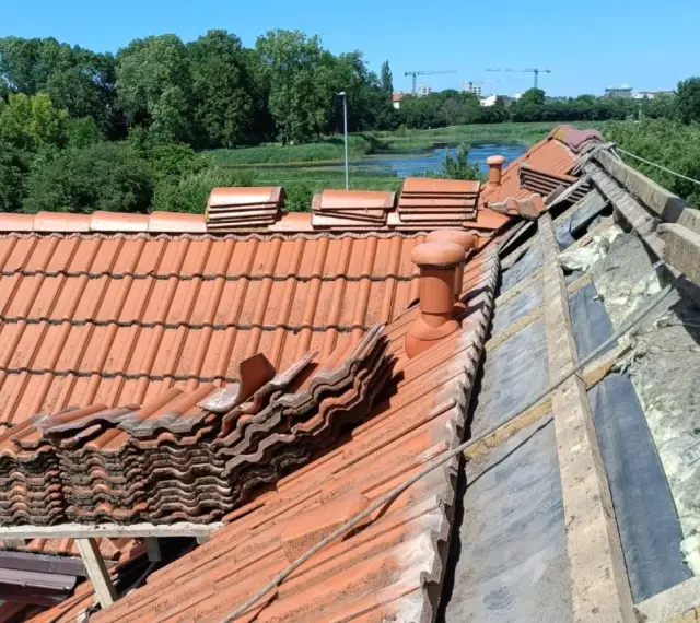 Trwa wymiana dachu. Stare dachówki są ułożone w stosy, a pod nimi widać izolację i drewniane elementy konstrukcji.